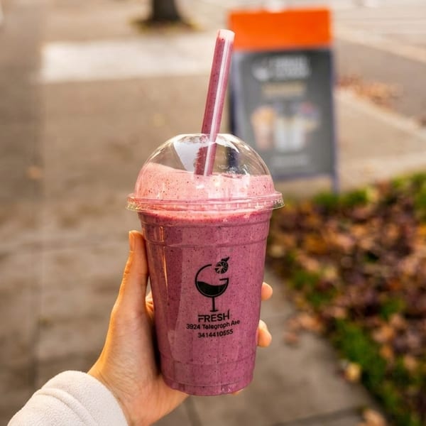 Hand holding a Fresh smoothie with a straw, outdoors on a sidewalk with foliage.