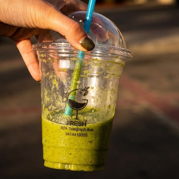 Hand holding a partially consumed green smoothie from "Fresh" with a blue straw.