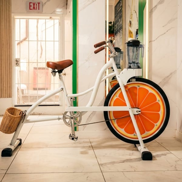White stationary bike with an orange slice wheel and a blender attached, on a marble floor.
